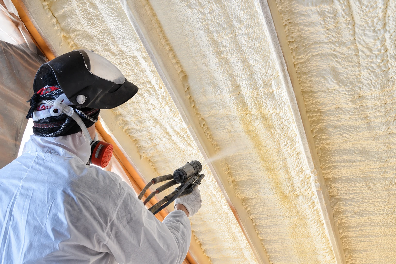 A worker in protective gear sprays expanding foam insulation between exposed roof rafters.