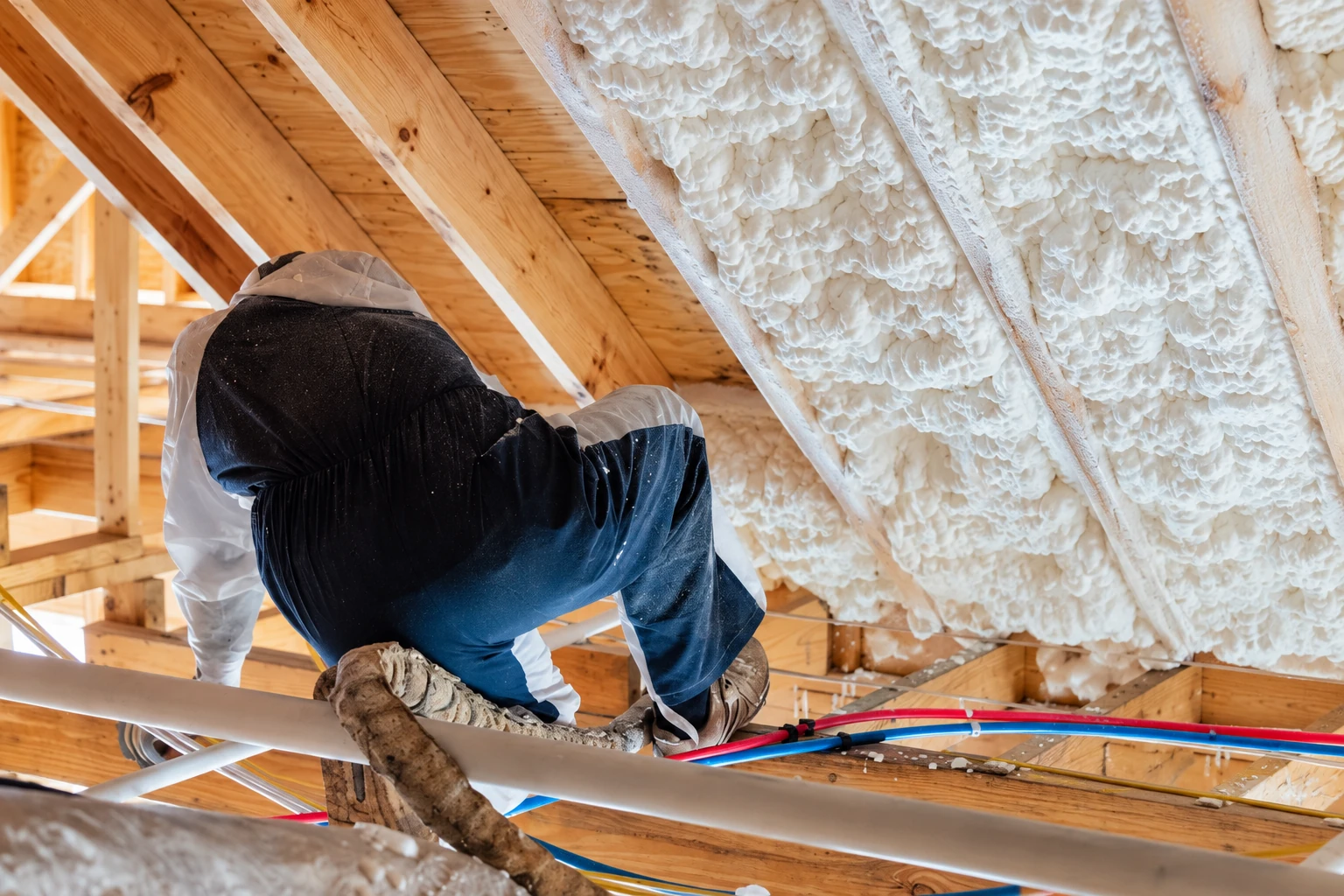 A worker in protective clothing applies spray foam to the underside of a wooden roof in an attic.