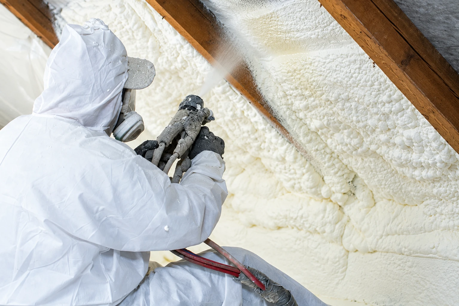 A worker in protective coveralls sprays white foam insulation onto the underside of a wooden roof.