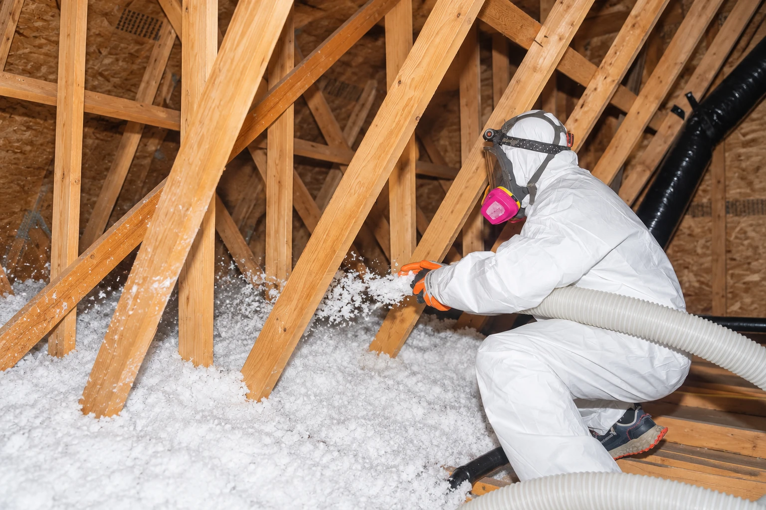 A worker in protective coveralls and a respirator blows loose-fill insulation into an attic using a flexible hose.
